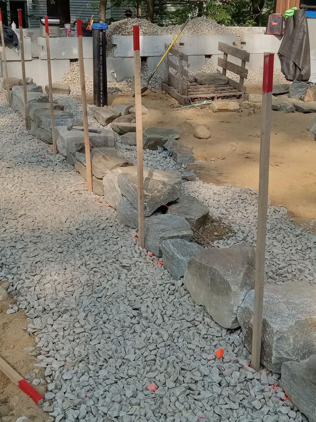 Large stones being set along a string line on a bed of crushed stone, with stakes marking the edge